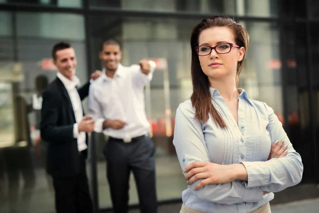 A woman standing in front of male coworkers who are making inappropriate comments about her. If you're experiencing illegal sexual harassment at work we recommend contacting a sexual harassment lawyer for guidance. 