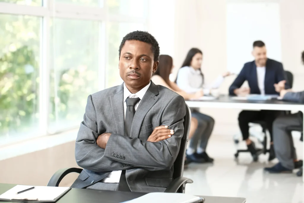 A black man sitting alone while his coworkers talk together in the background. If you're experiencing discrimination at work a discrimination lawyer in Austin can help.