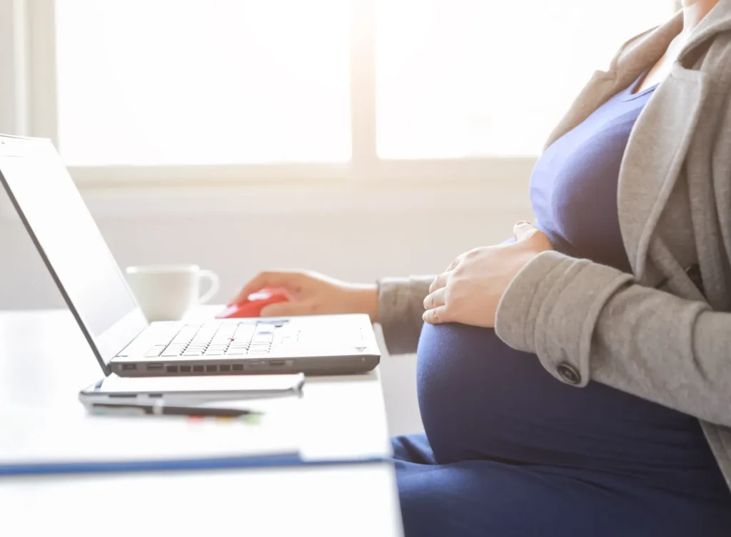 A pregnant woman hosting her belly and sitting at a desk working on her laptop.