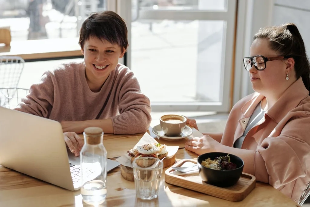 A woman smiling and working with another woman with down syndrome at a table looking at a laptop.
