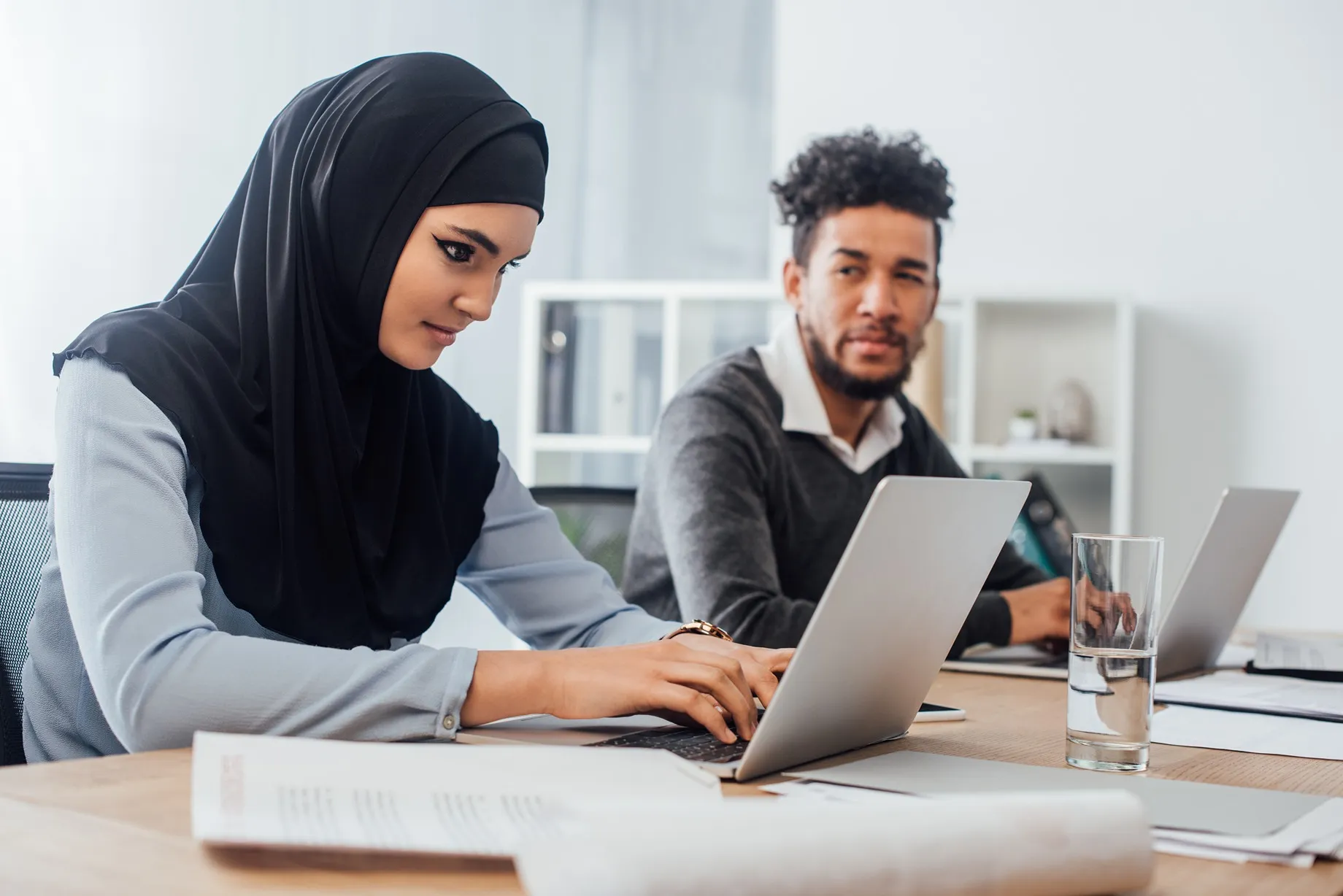 A female employee wearing a hijab and a male employee working at a desk together.