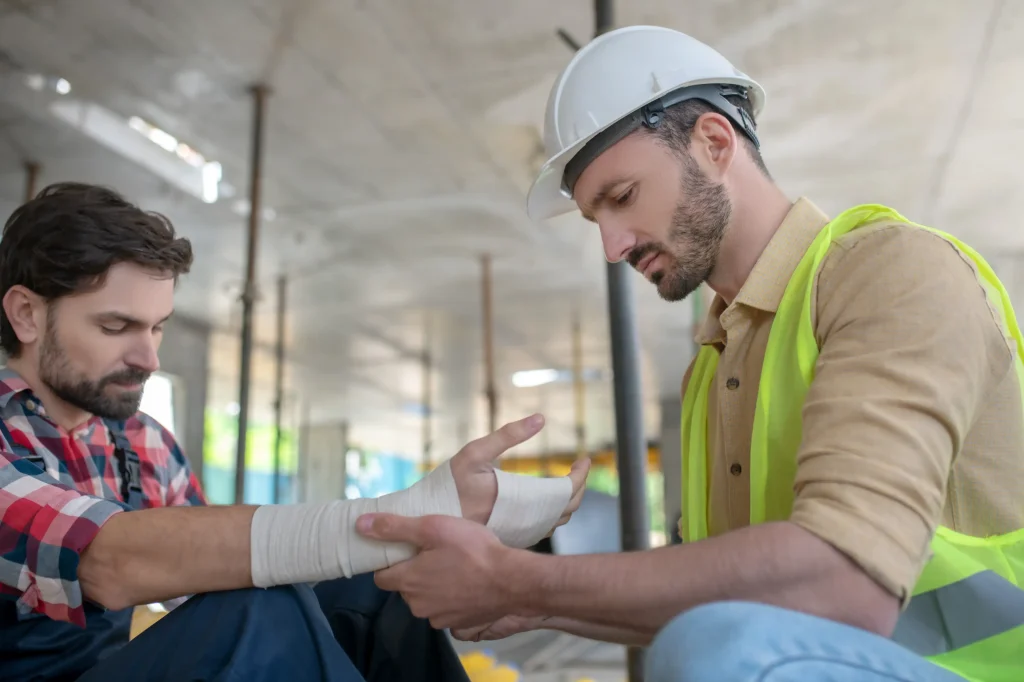An injured worker getting his wrist wrapped by his supervisor in a hard hat and high visability vest.