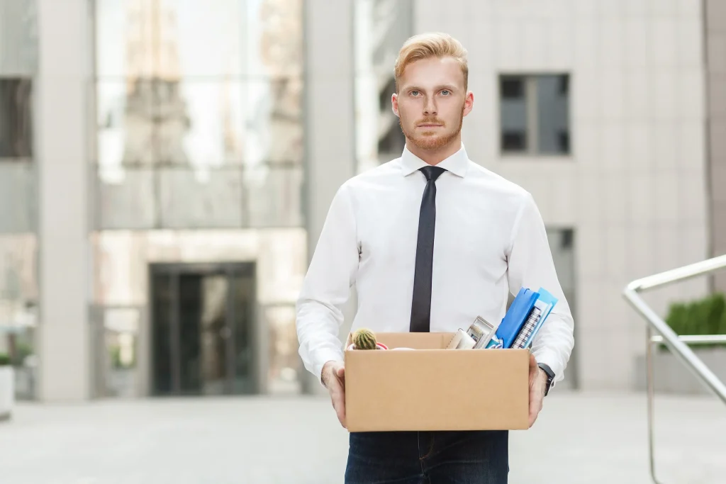 Man in business attire holding box of office supplies after being laid off, standing outside office building.